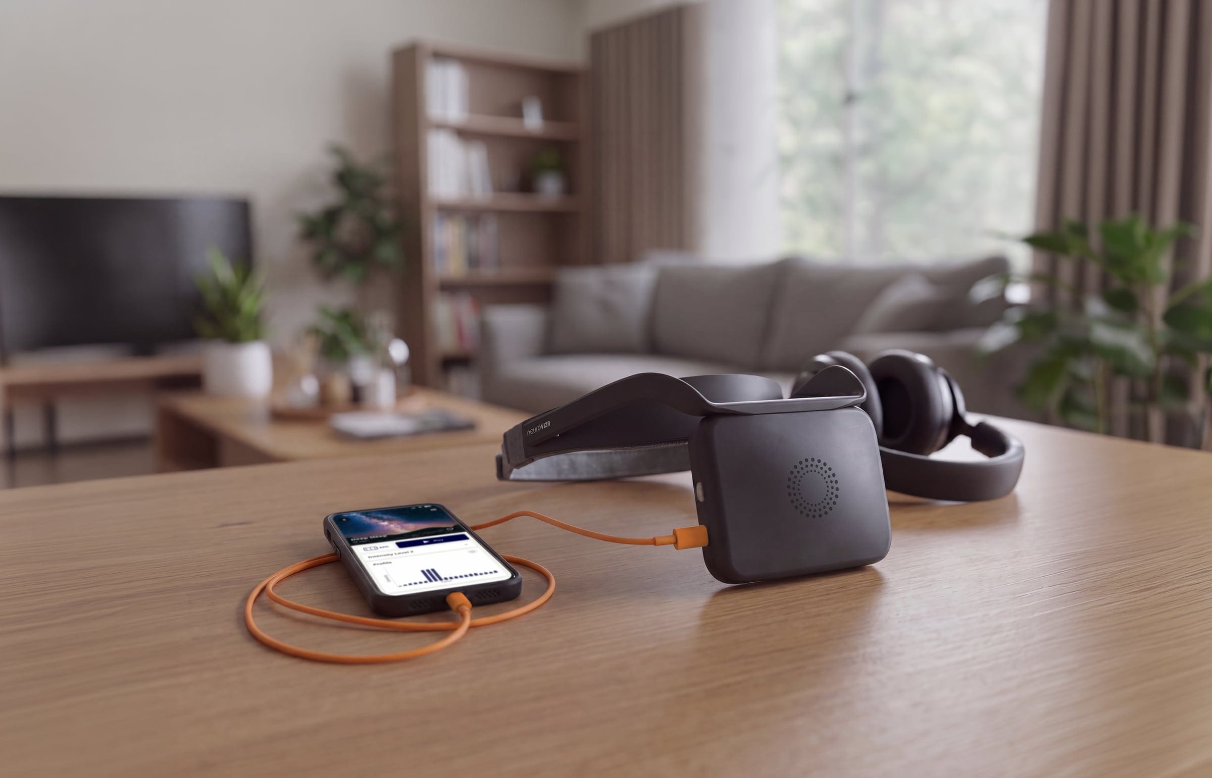 neuroVIZR LITE with headgear on a wooden table in a living room, connected to a smartphone by an orange cable, headphones behind it